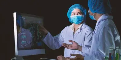 Two scientists wearing protective gear and masks, discussing and analyzing data displayed on a computer screen in a laboratory setting.