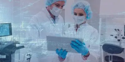 Two technicians in a laboratory wearing protective gloves, hair nets, face masks and lab coats looking at a tablet device