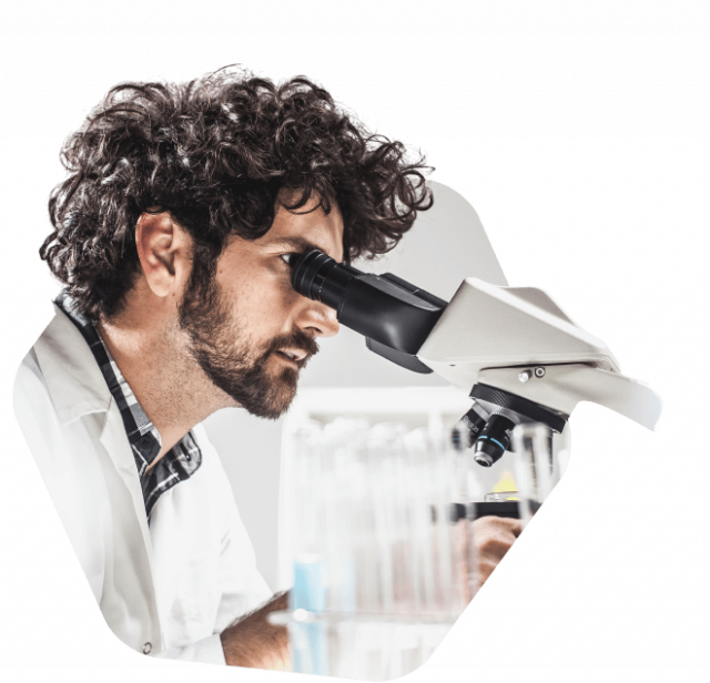 A male scientist with curly hair and a lab coat examines a sample through a microscope in a laboratory.